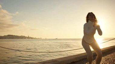 Atletik kadın şafak vakti nehir kenarında dinamik hareketlerle enerji veriyor, şehrin huzurlu güzelliğini kucaklıyor.