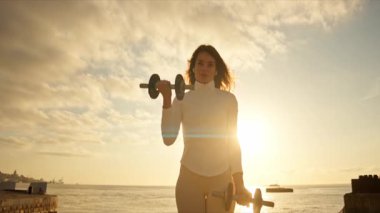 Fitness modeli iskelede gündoğumunda halterlerle egzersiz yapıyor.