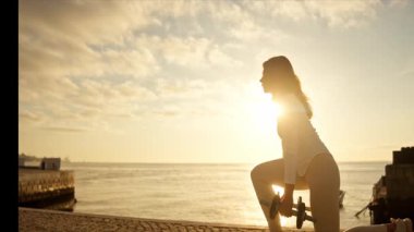 Dambıllarla egzersiz yapan, gün doğumunda deniz kenarında hamleler yapan genç bir kadın.