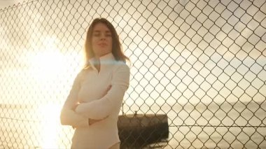 Gün batımında plajda çapraz kollu bir tel örgüyle duran genç bir sporcu.