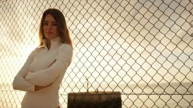 Kadın, arka planda deniz manzaralı ve gün batımlı tel örgülerin yanında dururken kendine güvenir.