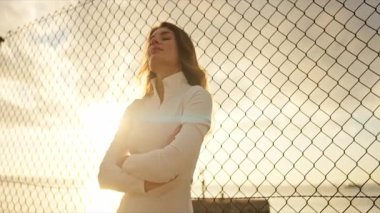 Genç bayan sporcu gün batımında kolları çapraz bir şekilde tel örgüye bağlı duruyor.