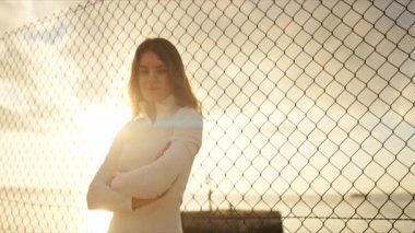 Çapraz kollarla poz veren genç bir sporcu tel örgüye karşı. Arka planda güneş denizin üzerinde batıyor.