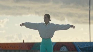 Genç bir kadın zarif bir şekilde dans hareketleri yapıyor kollarını açarak manzaralı bir günbatımı arka planına karşı, sükuneti ve odaklanmayı somutlaştırıyor.