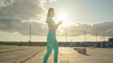 Spor giyimli genç bir kadın şehir ortamında günbatımında akıllı telefon kullanıyor.