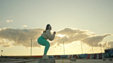 Kararlı sporcu kadın gün batımında kentsel çevrede beton blokta çömelme egzersizi yapıyor.