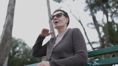 Orta yaşlı, gözlüklü bir kadın, uzun ağaçlarla çevrili bir bankta otururken el kol hareketi yapıp sohbet ederken görülüyor.