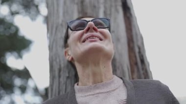 Yaşlı kadın, parkta bir ağacın yanında farklı duygular ve duygular ifade eden gözlük takıyor.