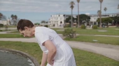 Orta yaşlı bir kadın, parktaki çeşmenin üzerinden eğiliyor, ellerinde su topluyor, huzurlu bir yaz günü sergiliyor.