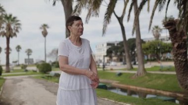 Yaşlı bir kadın parkta yürümekten zevk alıyor, el kol hareketi yapıyor ve konuşuyor.