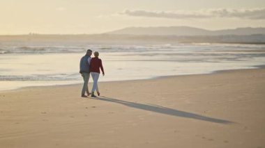 Emekli çift, altın bir günbatımında kumsalda romantik bir yürüyüş yapıyor.