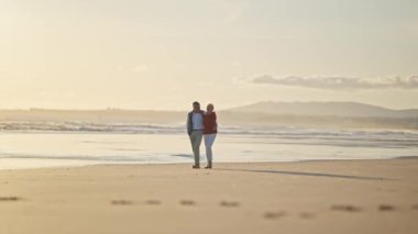 Emekli çift, altın bir günbatımında kumsalda romantik bir yürüyüş yapıyor.