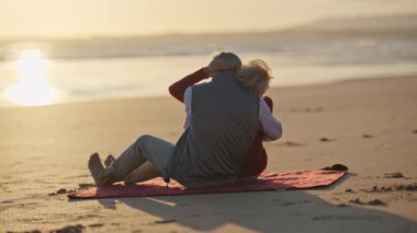 Sevecen yaşlı çift canlı bir günbatımında plaj battaniyesinde huzurlu bir anın tadını çıkarıyor.
