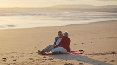 Emekli çift gün batımında battaniyede dinleniyor, okyanus manzarasının tadını çıkarıyor.