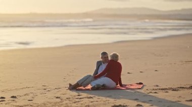 Yaşlı çift kırmızı battaniyede dinleniyor, gün batımında okyanusun kenarında samimi anlar paylaşıyorlar.