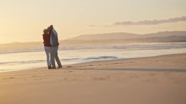 Emekli çift, altın bir günbatımında kumsalda romantik bir dansın tadını çıkarıyor.