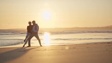 Emekli çift, altın bir günbatımında kumsalda romantik bir dansın tadını çıkarıyor.