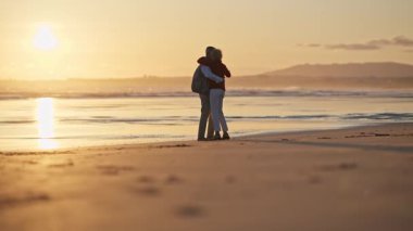 Romantik yaşlı çift gün batımında kumsalda huzurlu bir anın tadını çıkarıyor, kucaklaşıyor ve ufku işaret ediyor.