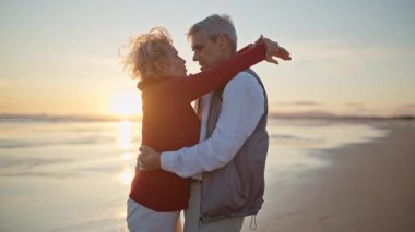 Yaşlı çift gün batımında sahilde romantik bir anın tadını çıkarıyor.