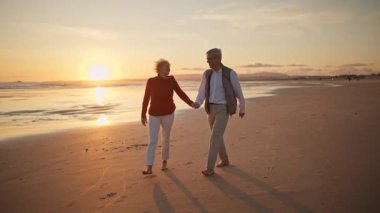 Emekli çift güzel bir gün batımında sahilde romantik bir yürüyüş yapıyor.