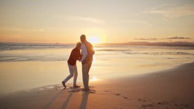 Emekli çift, güzel bir günbatımında sahilde romantik bir dansın tadını çıkarıyor.