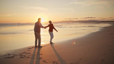 Kıdemli çift el ele tutuşuyor, yürüyor ve gün batımında sakin bir kumsalda kucaklaşıyorlar.