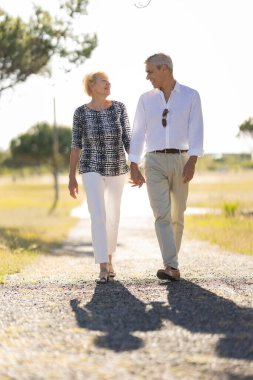 Emekli çift güneşli bir günün tadını çıkarıyor. El ele tutuşarak bir yolda yürüyorlar.