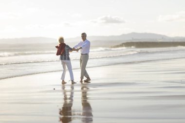 Kaygısız yaşlı çift gün batımında sahilde el ele tutuşup dans ediyor, emekliliğin tadını çıkarıyorlar.