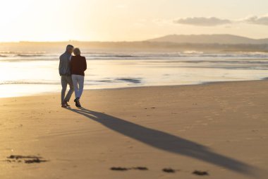 Yaşlı çift, altın bir günbatımında sahilde romantik bir yürüyüş yaparak emekliliklerini kutluyor.