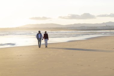 Yaşlı çift gün batımında sahilde romantik bir yürüyüş yapıyor.
