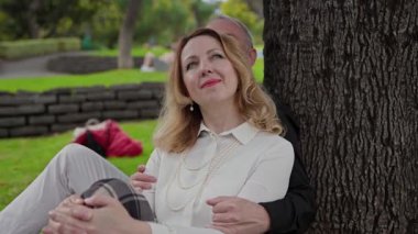 Romantik, olgun bir çift, canlı bir şehir parkındaki bir ağacın altında huzurlu dakikaların tadını çıkarıyor.
