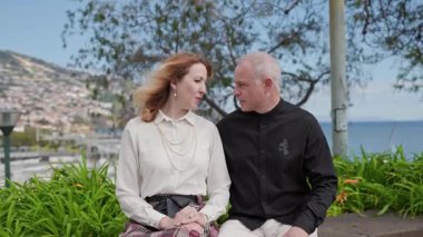 Olgun bir çift bankta oturuyor, konuşuyor ve Portekiz, Madeira Adası 'ndaki funchal limanının manzarasını seyrediyor.