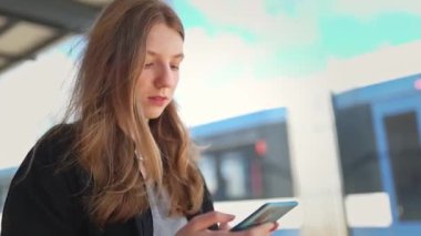 Sarışın kadın tren istasyonunda beklerken akıllı telefon kullanıyor.