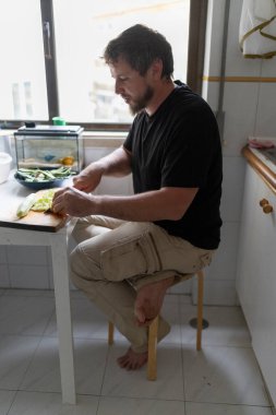 Sakallı adam taburede çıplak ayakla oturmuş titizlikle mutfakta sebzeleri küçük teraryumla doğruyor.