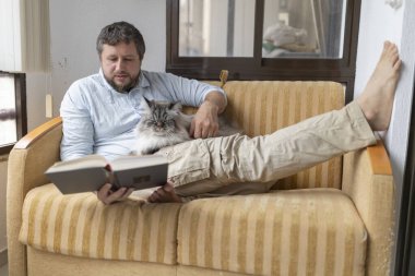 Evdeki rahat bir kanepede, tüylü kedisi kucağında huzur içinde uyurken kitap okumaktan zevk alan bir adam.