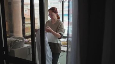 Kızıl saçlı kadın evinin balkonuna temiz beyaz elbiseler asıyor.