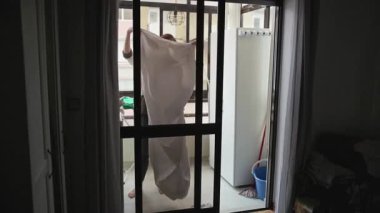Ev hanımı, apartmanın balkonundaki çamaşır ipine yeni yıkanmış beyaz çarşafları asıyor.
