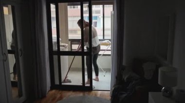 Kadın hizmetçi rutin temizlik işi yapıyor, el süpürgesi kullanarak balkon zeminini süpürüyor, gündüz şehir yaşamında ev hijyenini koruyor.
