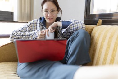 Kanepede dizüstü bilgisayar kullanan, çay içen, evden çalışan bir kadın.