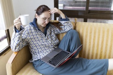 Kanepede rahat bir şekilde oturan bir kadın, dizüstü bilgisayarla çalışırken bir fincan kahvenin tadını çıkarıyor.