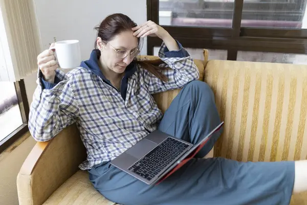 Kanepede rahat bir şekilde oturan bir kadın, dizüstü bilgisayarla çalışırken bir fincan kahvenin tadını çıkarıyor.