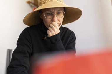 Hasır şapka takan ve gözlük takan kadın sanatçı elinde boya fırçası tutarken konsantre oluyor.