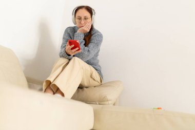 Kablosuz kulaklık takan ve evdeki koltukta otururken akıllı telefonunu karıştıran olgun bir kadın.