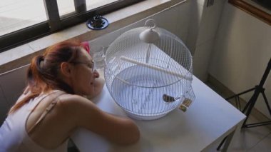 Kızıl saçlı kadın beyaz bir kafeste evcil kuşuyla konuşuyor.