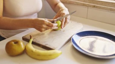 Kadın, bir muz ve başka bir elmanın yanında, tahta kesme tahtasındaki yeşil elmayı dilimliyor.