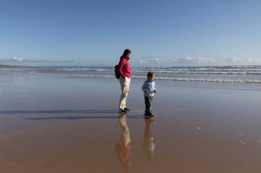 Anne ve oğlu, dalgaların çekildiği bir kumsalın ıslak kumlarında sakin bir şekilde yürüyorlar. Yansımaları sığ suda görülebiliyor.