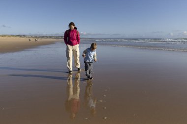 Anne ve oğlu güneşli bir günde ıslak kumda sakin bir şekilde yürüyorlar.