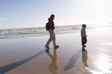 Anne ve oğul, güneş batarken kumların üzerinde uzun gölgeler oluşturarak rahatça geziniyorlar.