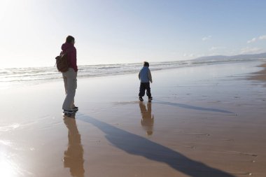 Anne ve oğlu gün batımında kumsalda huzur içinde yürüyüp ıslak kumda uzun gölgeler yaratıyorlar.
