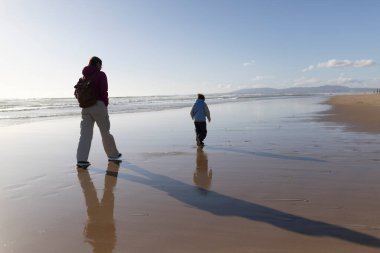 Anne ve oğlu, güneş batarken sahil boyunca huzur içinde yürüyüp ıslak sahilde uzun gölgeler yaratıyorlar.
